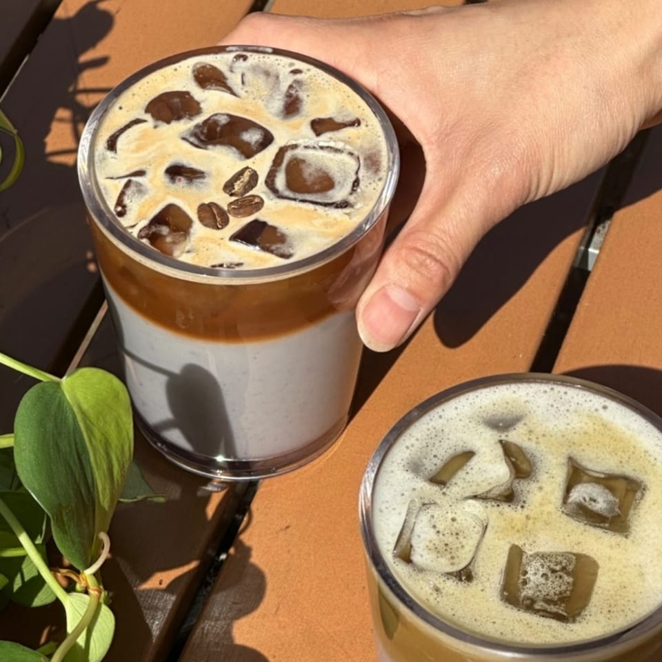 A hand holds a glass of iced coffee with coffee beans, next to another iced coffee.