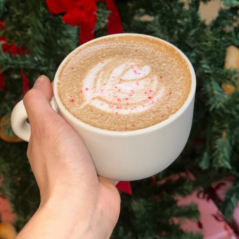 Hand holding a white mug with latte art and red sprinkles, against a festive green background.