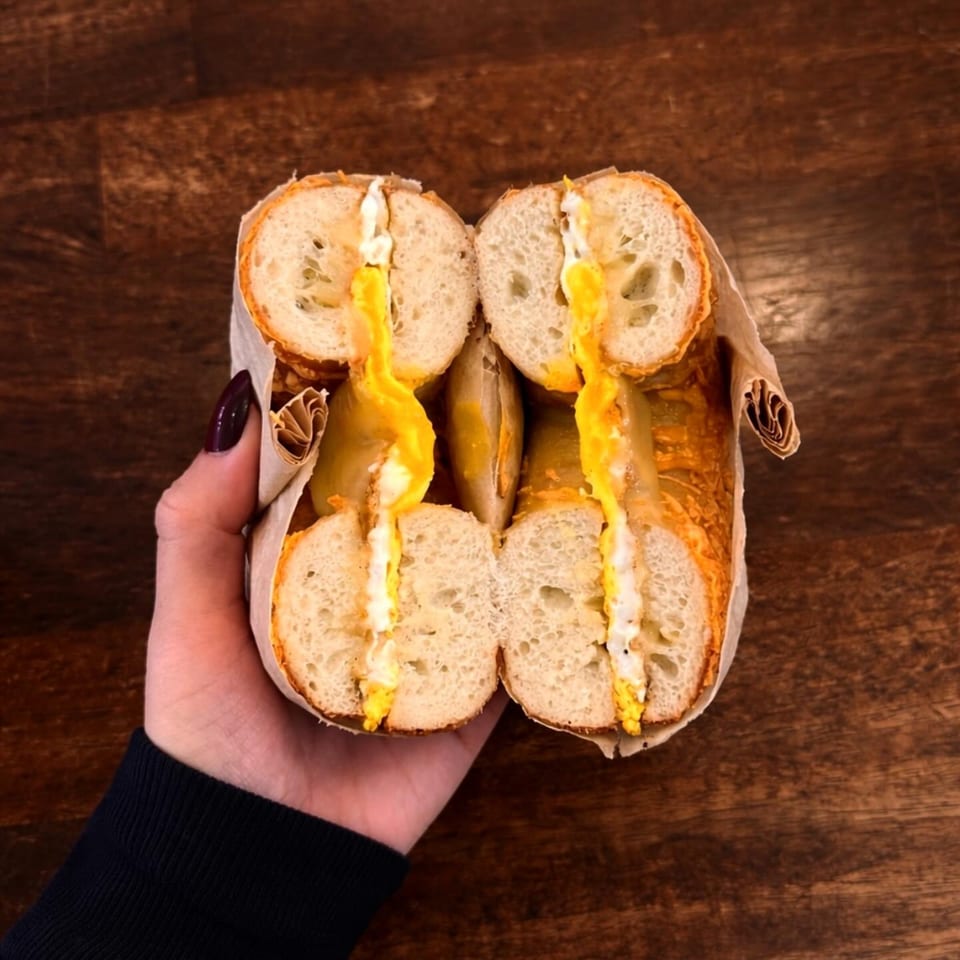 A hand holds a bagel sandwich cut in half, revealing melted cheese and egg inside.